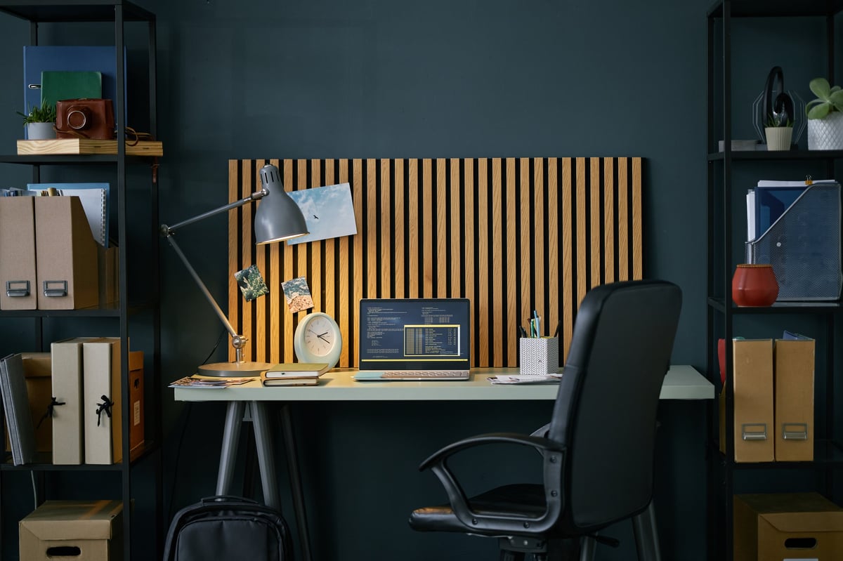 A warm, well-lit study workspace with an open laptop on a desk, a brass lamp, books and plants on surrounding shelves
