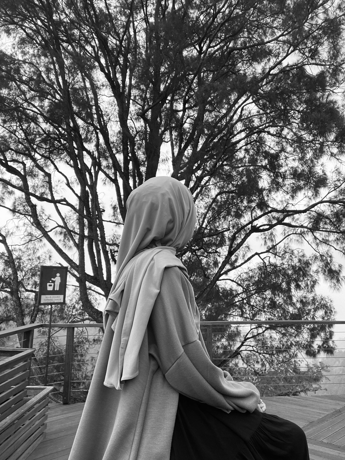 Woman wearing headscarf sitting on wooden deck overlooking large trees in black and white photo