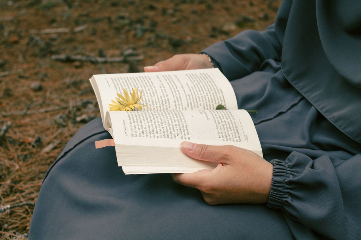 Person in dark jacket holding an open book with a yellow flower pressed between pages, sitting on the ground