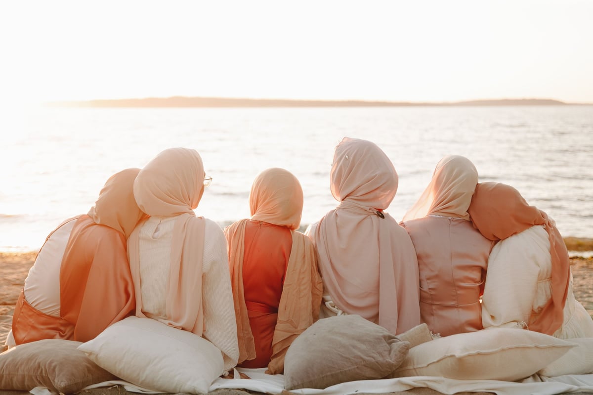 Group of women wearing hijabs sitting together on beach at sunset, viewed from behind