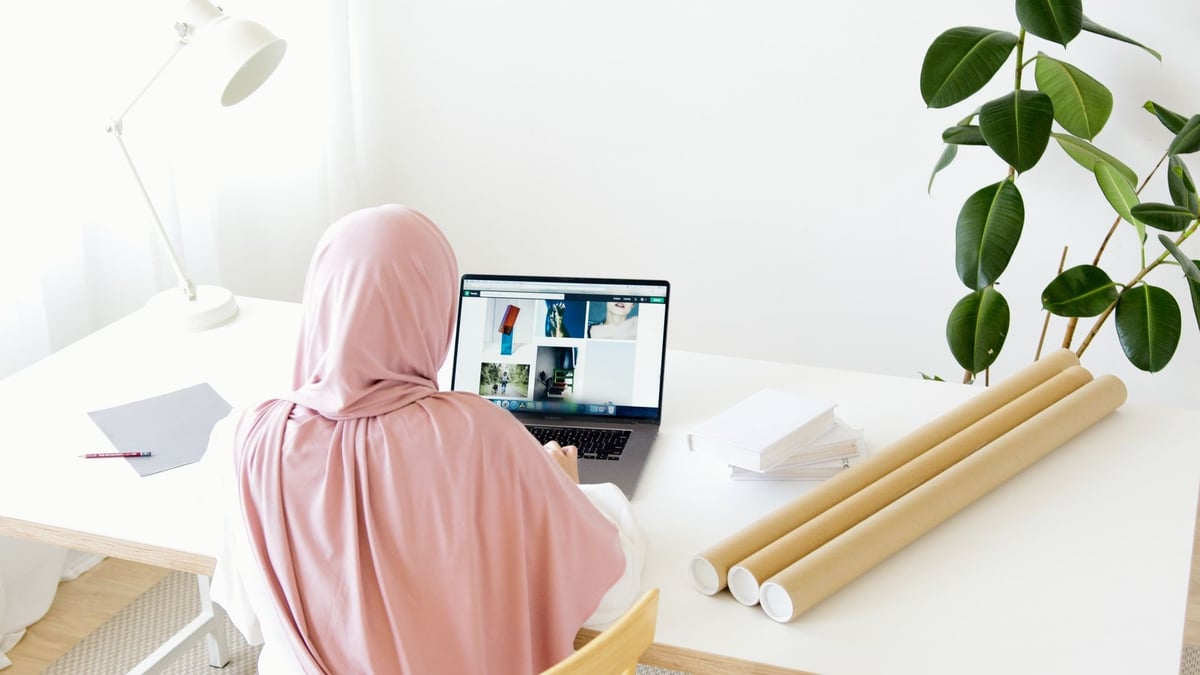 Woman wearing pink hijab working at white desk with laptop, surrounded by plants and desk accessories in bright office space.
