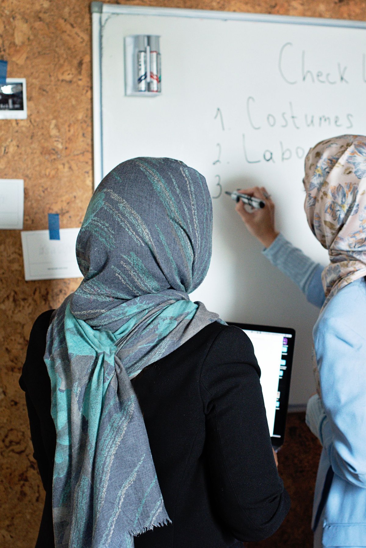 Person wearing a green headscarf writing on a whiteboard with a checklist, photographed from behind in front of a cork wall.