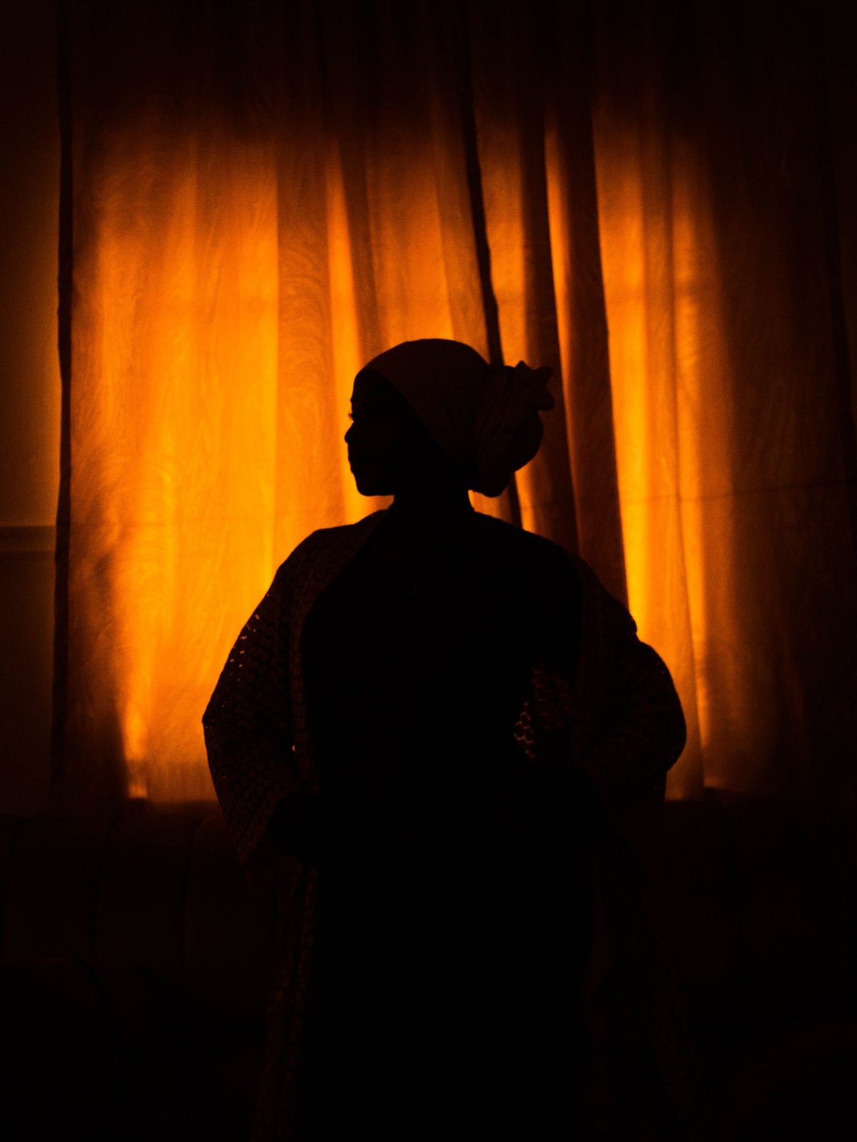 Silhouette of a person in a headscarf standing before a bright orange curtain, backlit by warm light